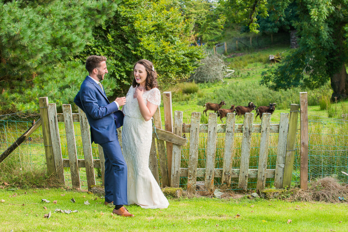 brunette bride wearing a vintage, beaded wedding dress with her groom wearing a blue suit and green, tweed tie standing in front of a wooden, picket fence with brown pygmy goats in the background