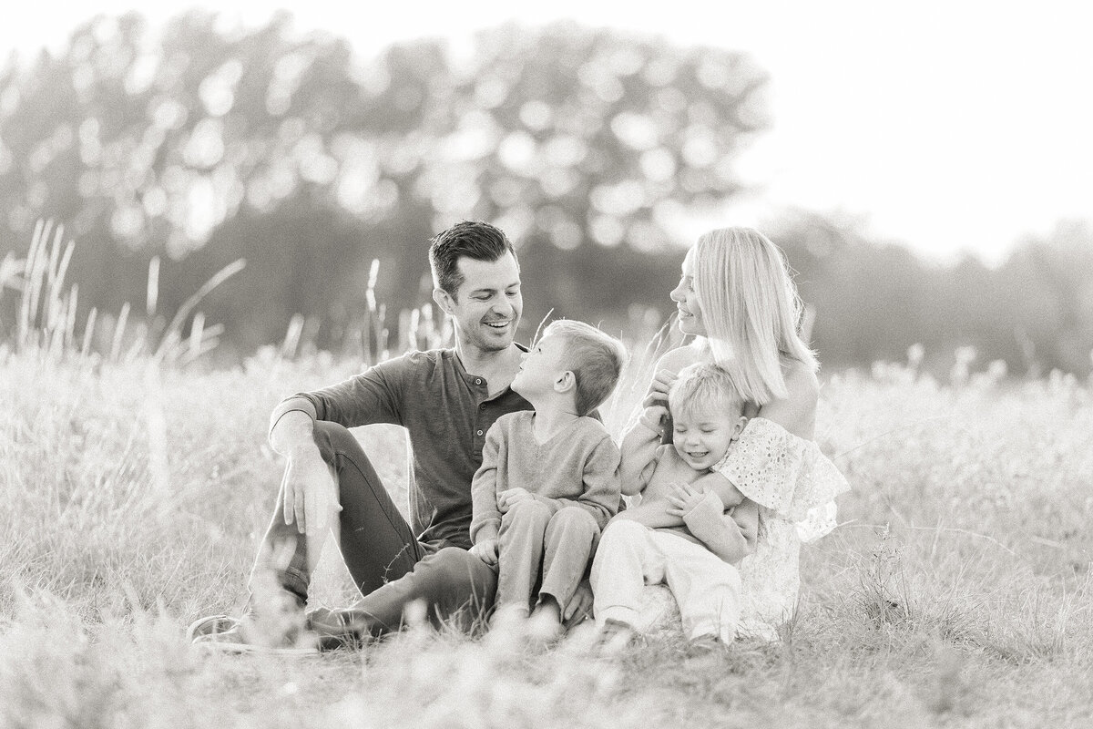 A black and white portrait of a young family of 4 taken while they are all sitting together in an open field at a local Dallas/Fort Worth park taken by a family photographer in Dallas Texas.