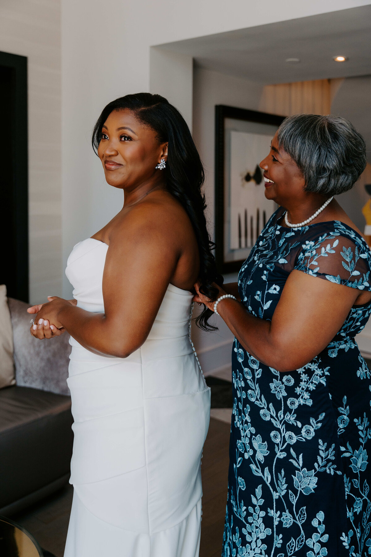 A bride in a white dress smiles warmly as an older woman in a blue floral gown adjusts her dress in a cozy room. The scene conveys love and joy.