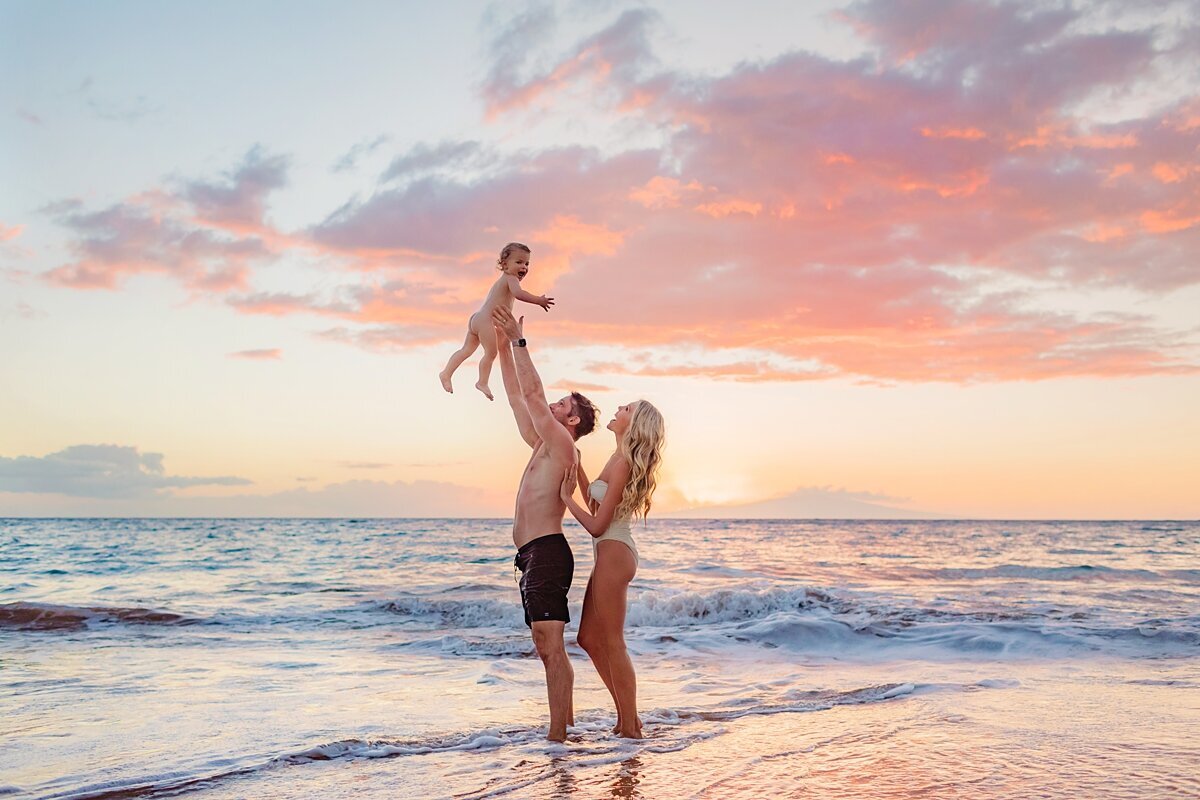 Blonde woman in white one piece swimwear holds her husband's shoulder and looks at her family at sunset on the beach in Maui