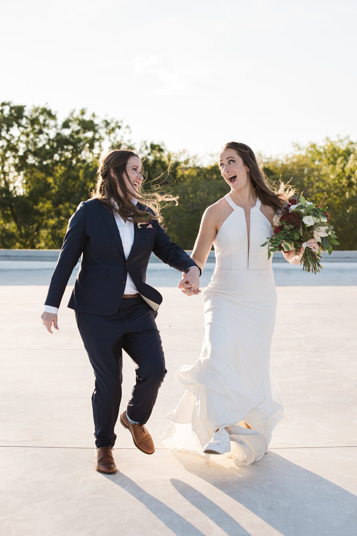 Two brides playfully walking and laughing on the roof of their venue after their wedding ceremony at the BRIK Venue in Fort Worth, Texas. The bride on the right is wearing a sleeveless, elegant, white dress and is holding a bouquet. The bride on the left is wearing a navy suit with a boutonniere.