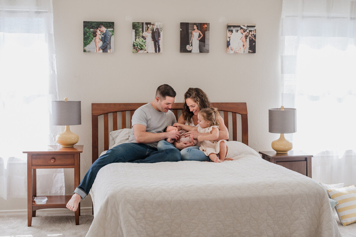 Pull-back shot of a family on their master bed admiring their new baby. Light streams in the windows on both sides of the bed.