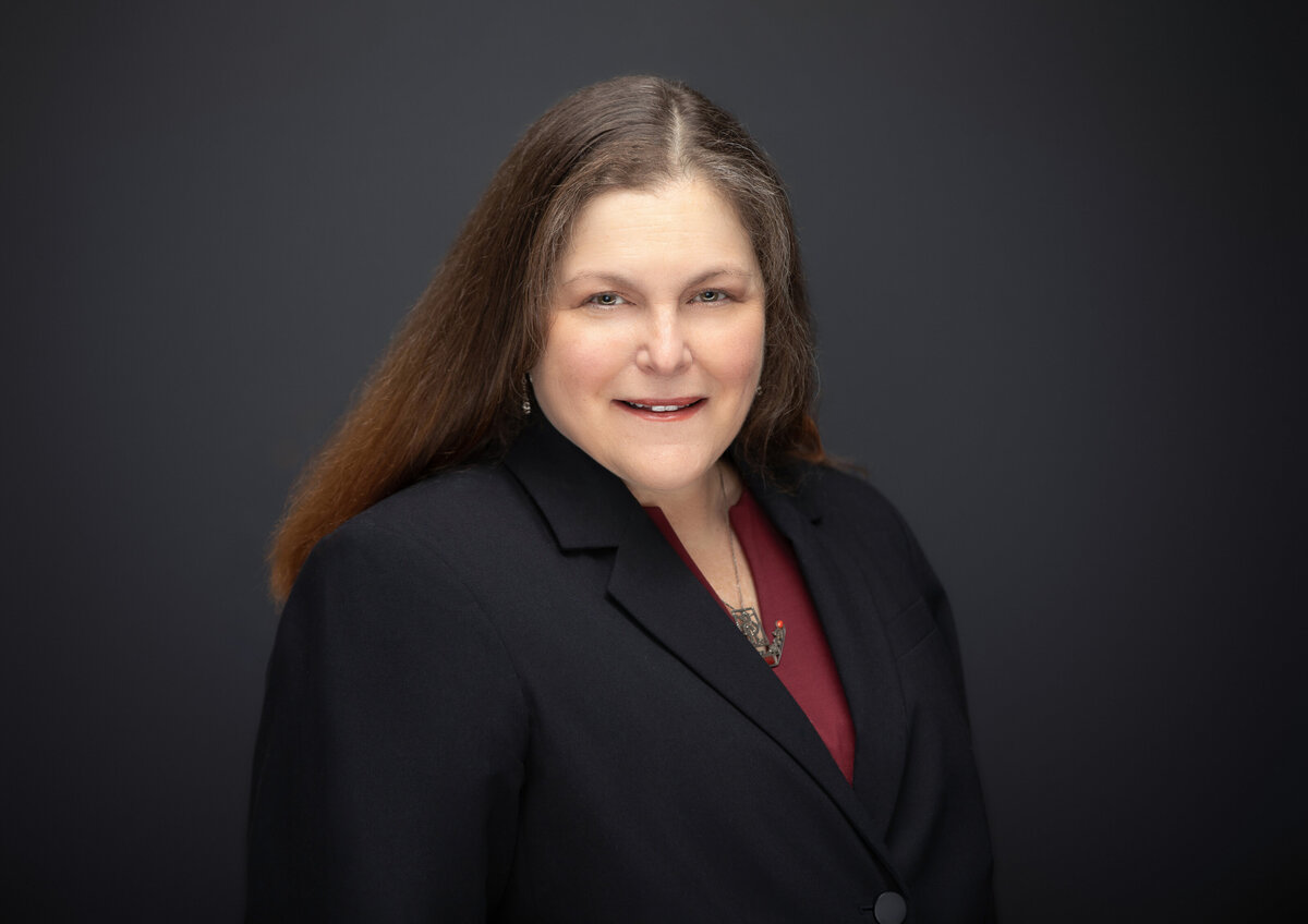 A woman female doctor medical professional in a black blazer and with long straight hair and maroon top poses for a professional headshot on a black backdrop for Janel Lee Photography studios in Cincinnati Ohio