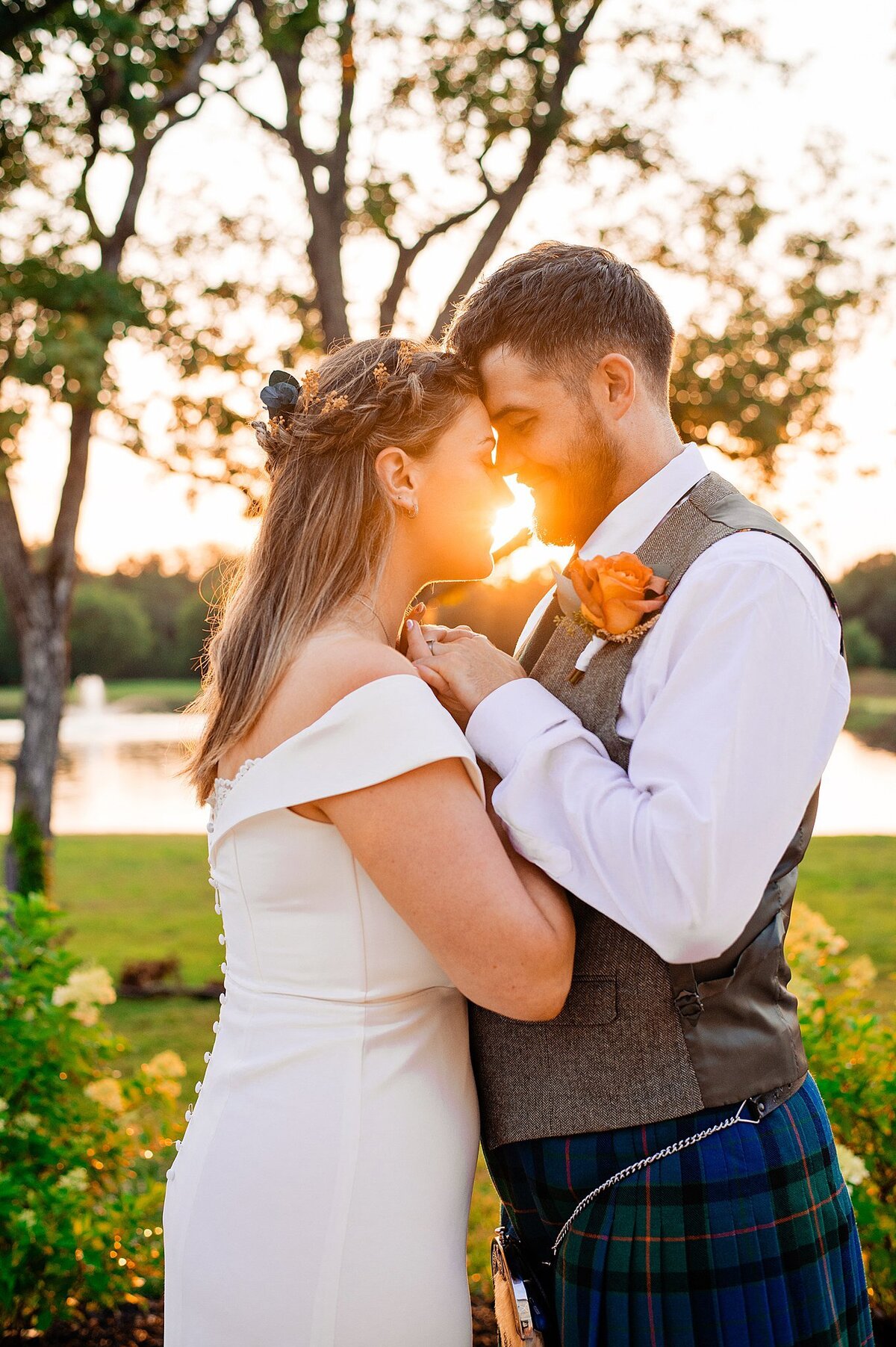 The bride, wearing a fitted gown with off the shoulder sleeves and flowers in her hair leans in to kiss the groom. The groom is wearing a gray vest, a white shirt and a blue and green kilt with sporran. He leans in to kiss the bride lakeside as the sun sets behind them at Sugarfoot.