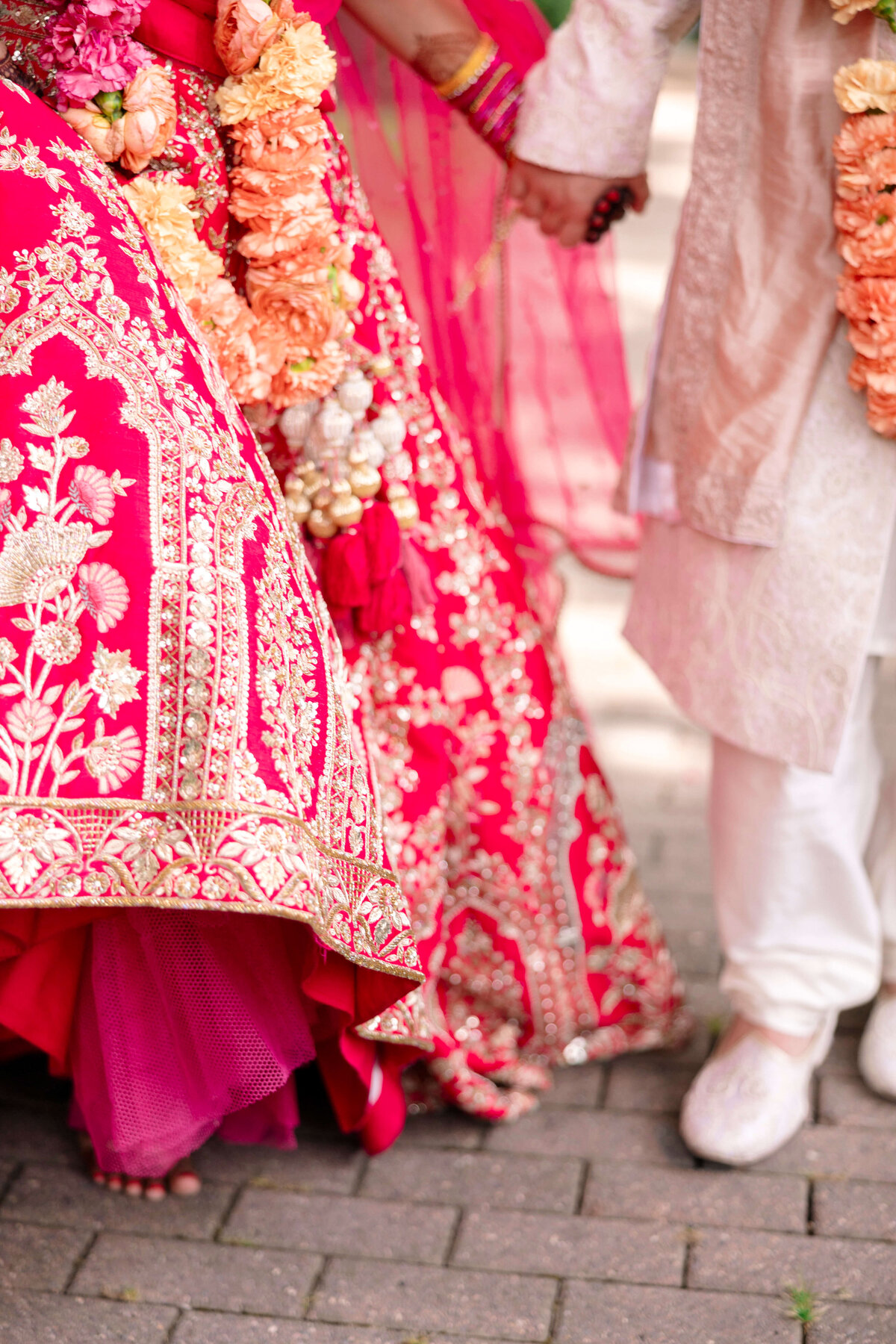 A couple wearing traditional Indian wedding attire holds hands. The brides vibrant red and gold embroidered lehenga is adorned with floral garlands, while the groom wears a cream-colored sherwani and white shoes.