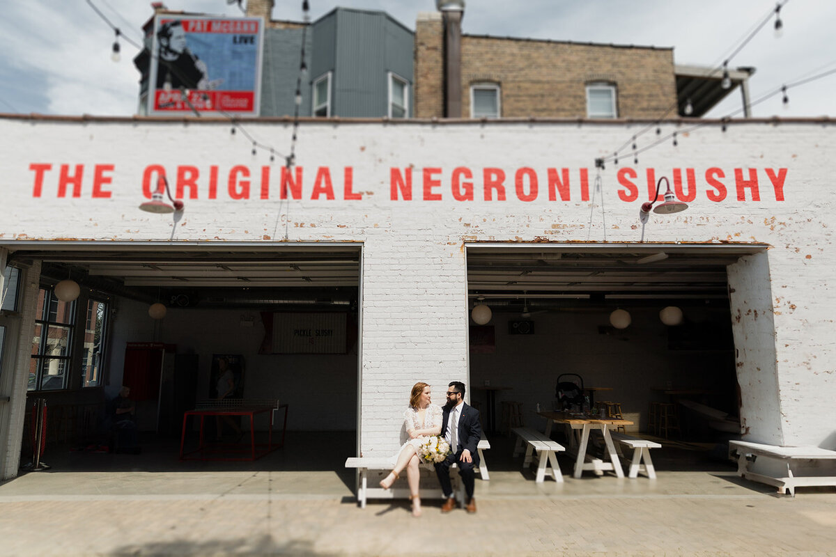 Elopement couple sits on a bench outside Parson's Chicken in Lincoln Park Chicago. It's very sunny, and the sign in red reads "The Original Negroni Slushy."