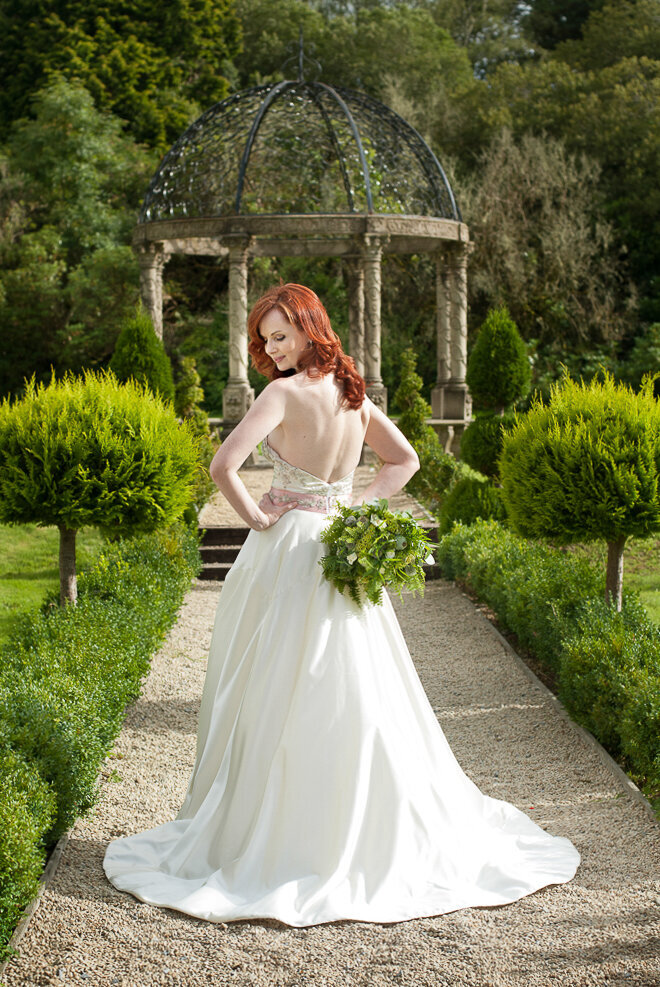 bride with long, silky, red hair wearing a ballroom style wedding dress with beaded bodice looking over her shoulder while holding a green wedding bouquet in the gardens of Ballyseede Castle, Kerry