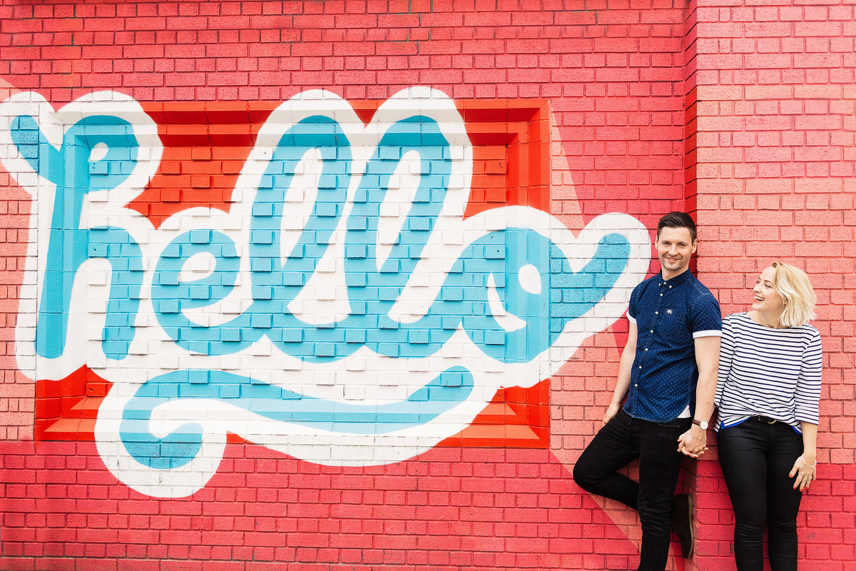 An engagement shoot photo of a couple in Leeds stood in front of street art that reads hello