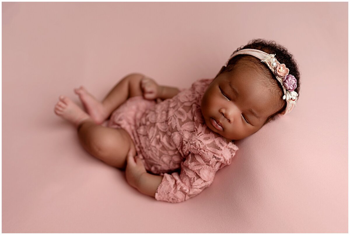 A newborn baby laying on a dreamy pink backdrop wearing a pink romper and pink headband.