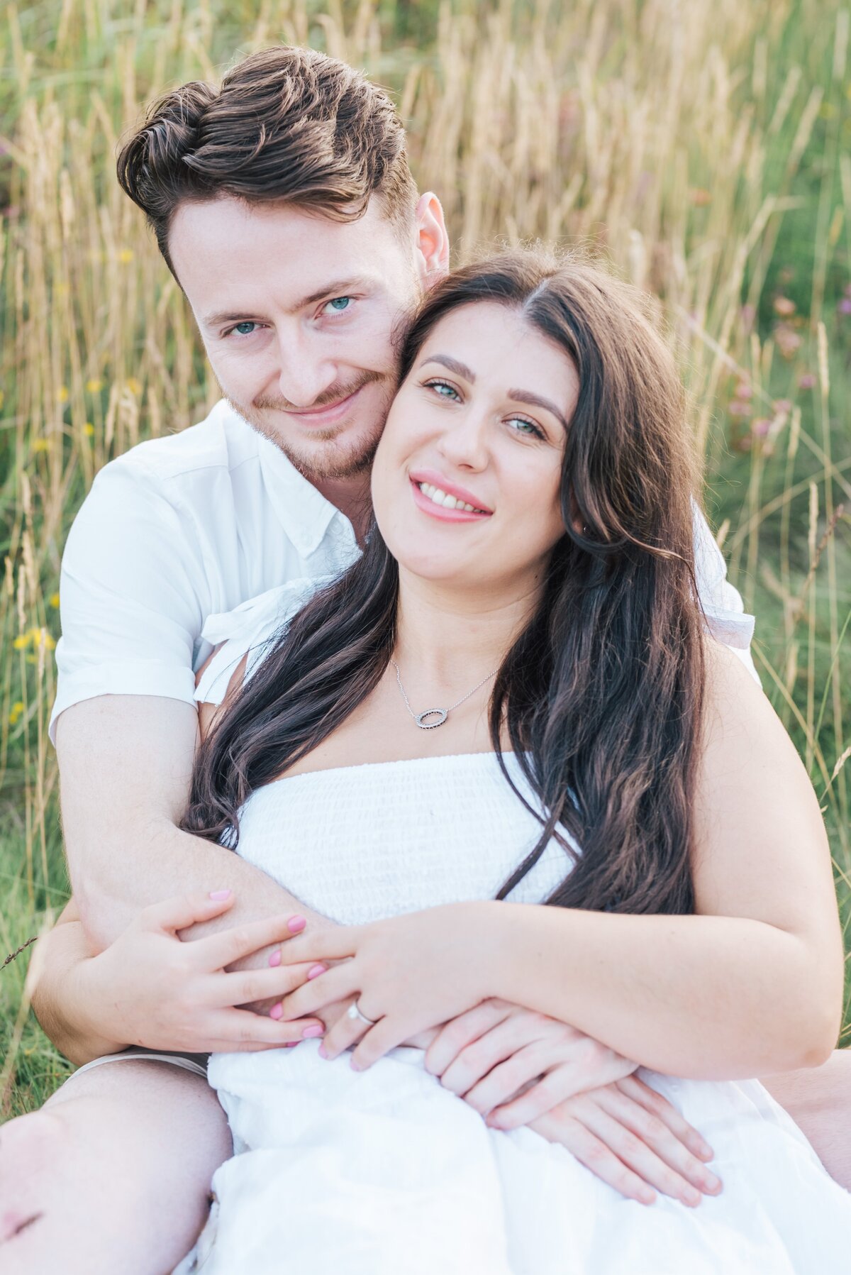 The couple are sitting in a field with long grass around them, the man is behind the lady and his arms around her, they are cuddling and smiling at the camera,