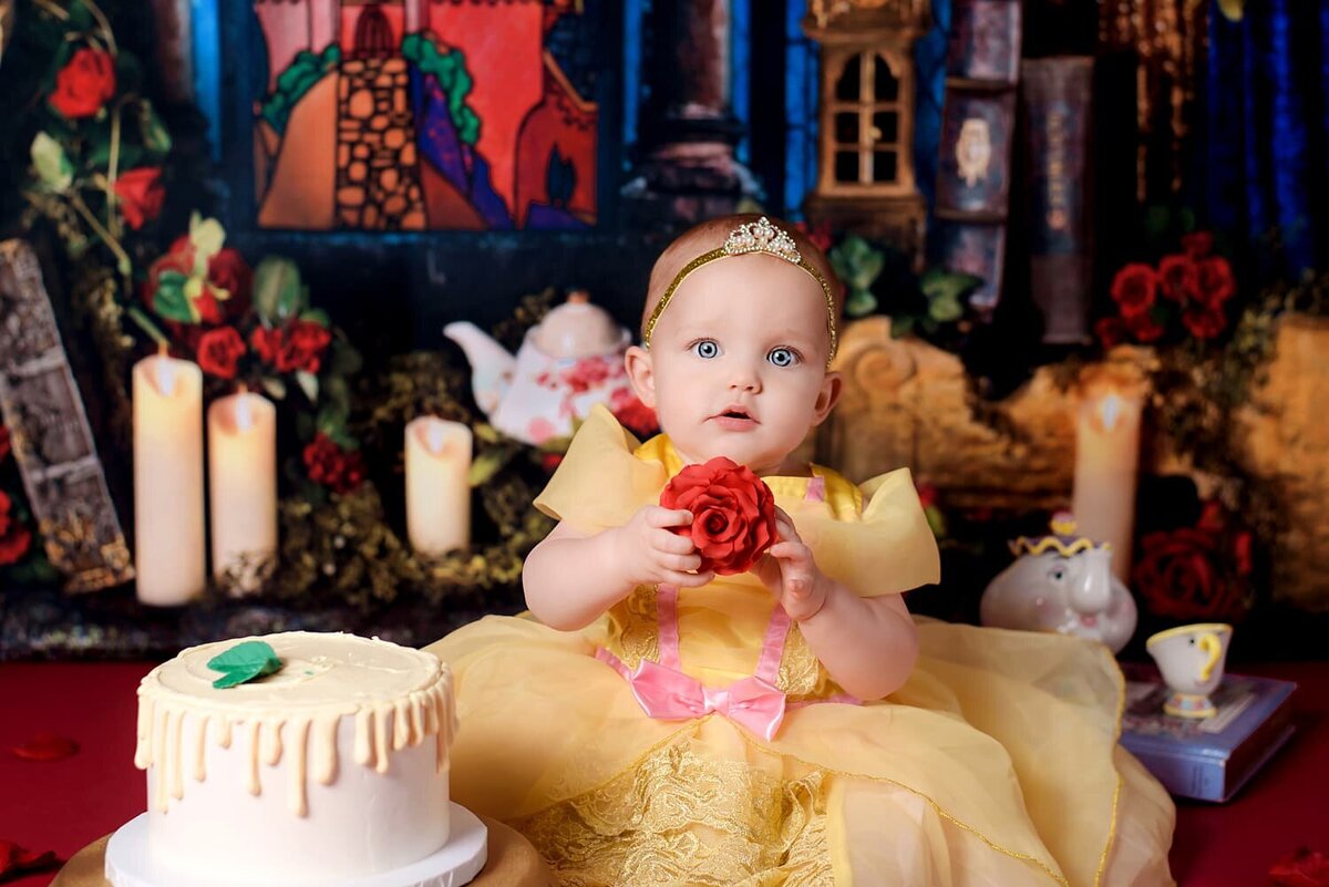 1 year old girl wearing yellow Belle dress holding a rose during cake smash.