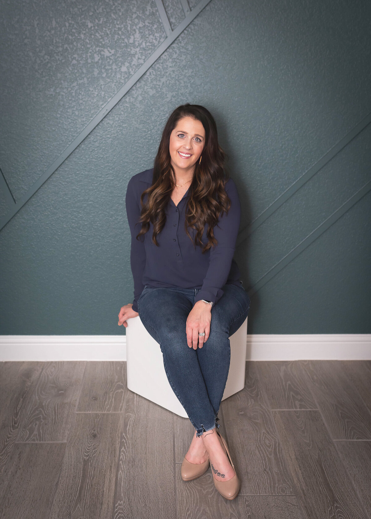 young woman in navy blue shirt jeans and tan high heels sitting on white posing block with a teal feature wall behind her