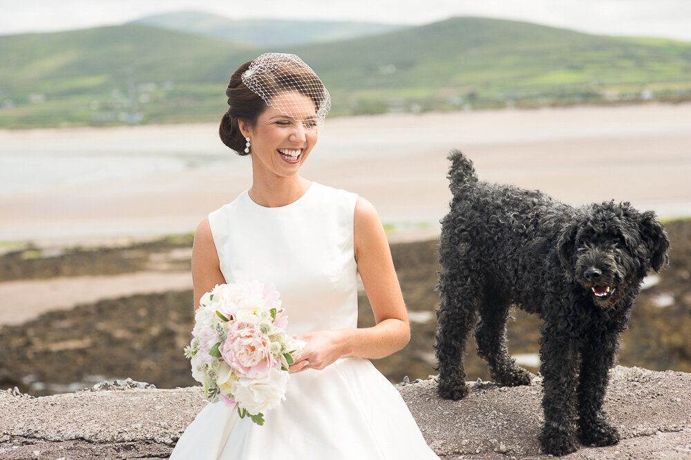 bride wearing a vintage, tea-length wedding dress and a net birdcage wedding veil, sitting on a wall overlooking the se with her black labradoodle while holding a bouquet of peonies