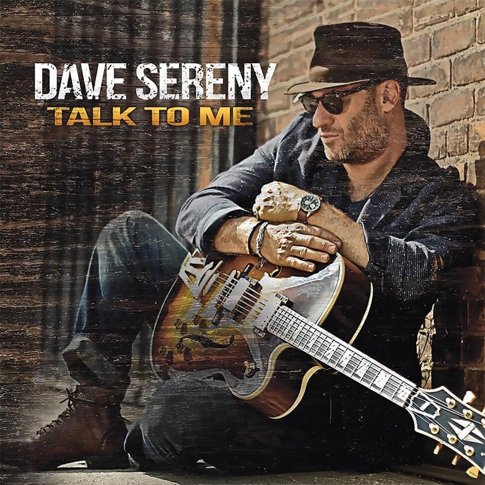 CD Cover Title Talk To Me Artist Dave Dereny sitting in front of gate and brick wall guitar in his lap wearing sunglasses