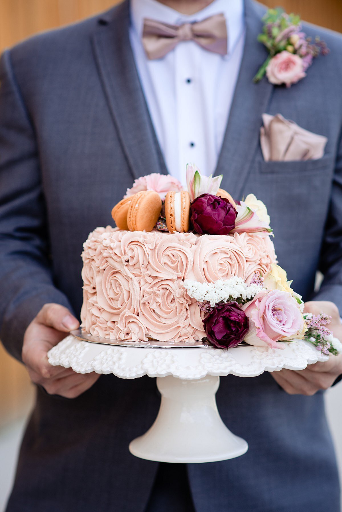 Blush one tier wedding cake with rosette icing. Topped with orange macarons, small peonies and roses. The cake is on a white footed cake stand being held by the groom who is wearing a dark blue suit, white shirt and light brown bowtie. He has a boutonniere of blush flowers to match his pocket square.