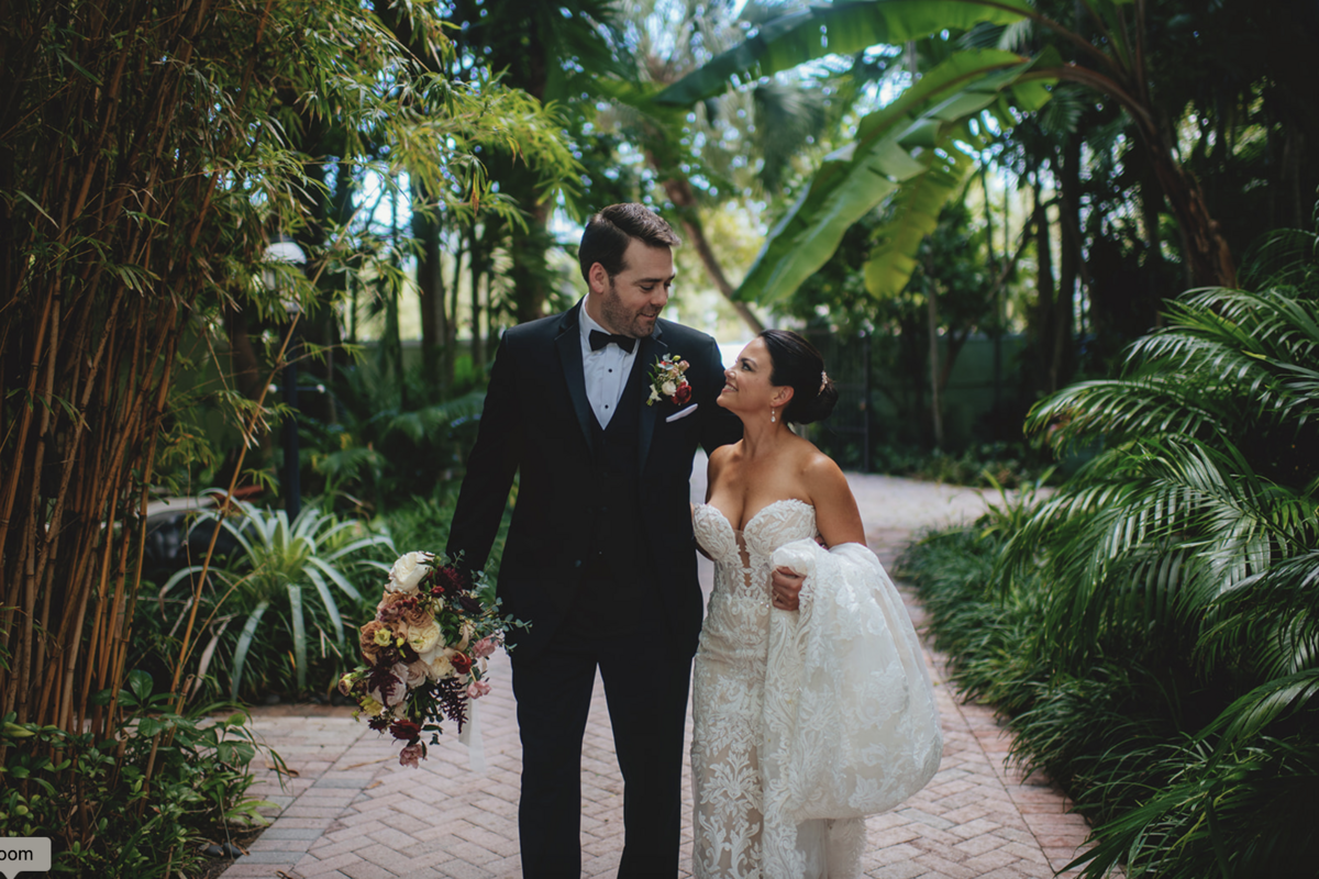 Elegant bride in lacy plunging dress smiles at groom on wedding day amidst lush greenery at Holly Blue.