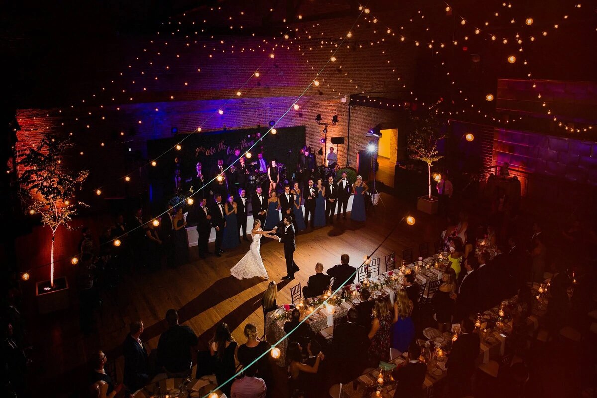 Color photo of a bride and groom in their first dance surrounded by guests under twinkling lights in a warm, inviting reception hall.