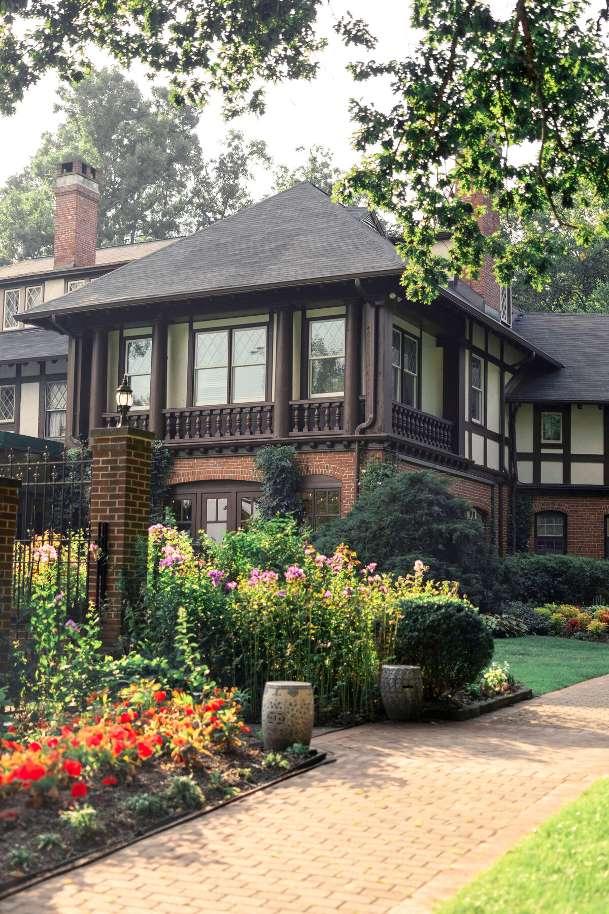 A charming Tudor-style house with a dark roof and exposed timber detailing. The front yard features a well-maintained garden with colorful flowers and a brick pathway leading to the entrance, surrounded by lush greenery and trees.