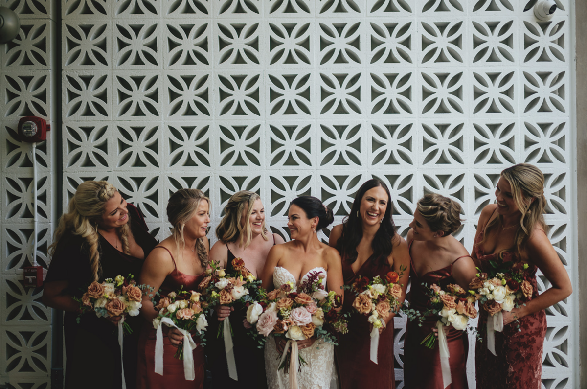 Bride in plunging gown smiles at six bridesmaids beside her in  garnet gowns outside of Fort Lauderdale wedding venue.