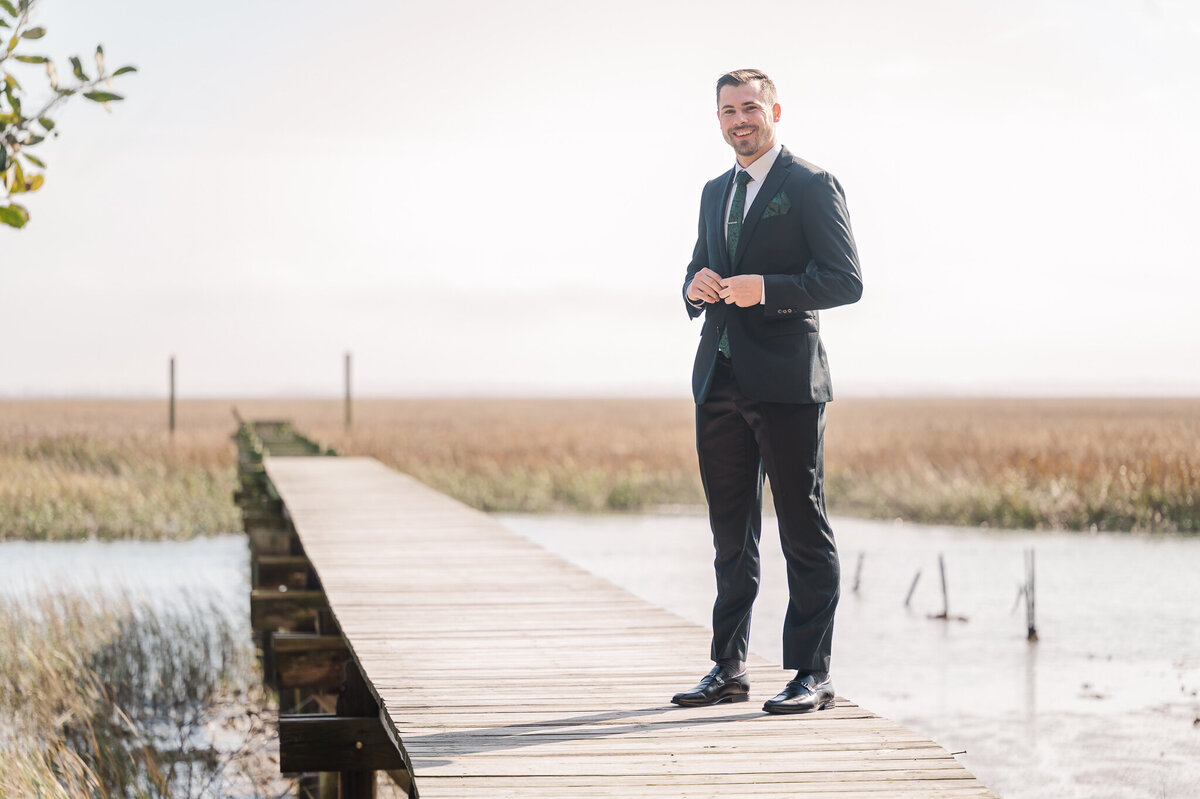 A groom on his wedding day buttoning his black suit jacket on a bridge overlooking the water enjoying his Raleigh wedding photography session by JoLynn Photography