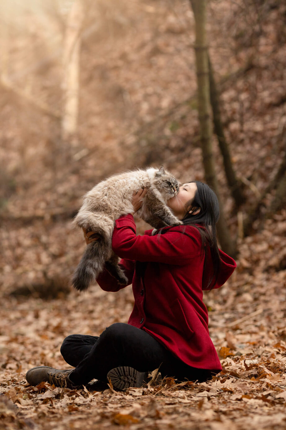 Woman in red coat sitting on a forest trail lifting her cat for a kiss