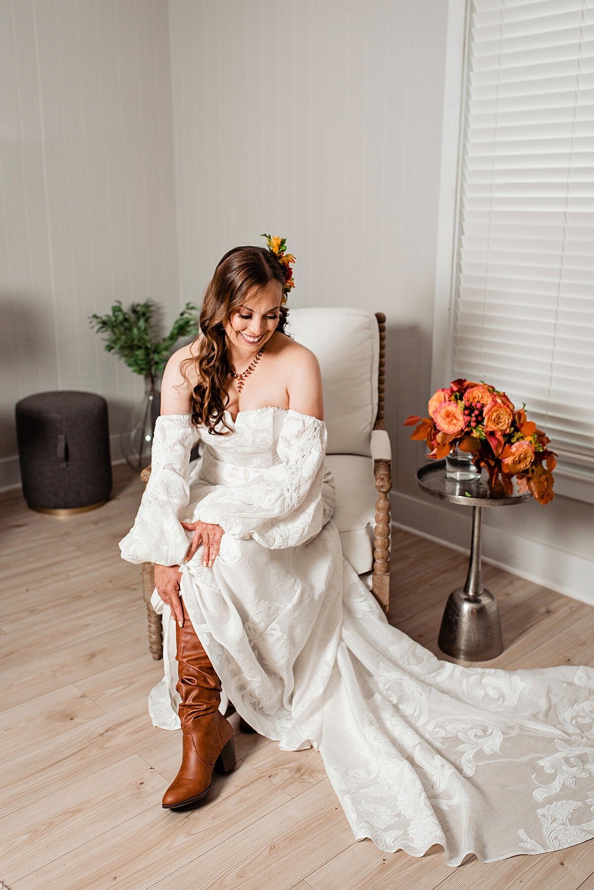 The boho bride is seated in a chair next to a silver table with her bouquet of orange flowers and dried autumn leaves. She is wearing a flowing sleeveless ivory gown with an empire waist and detachable sleeves. She is leaning down to put on knee high brown leather boots.