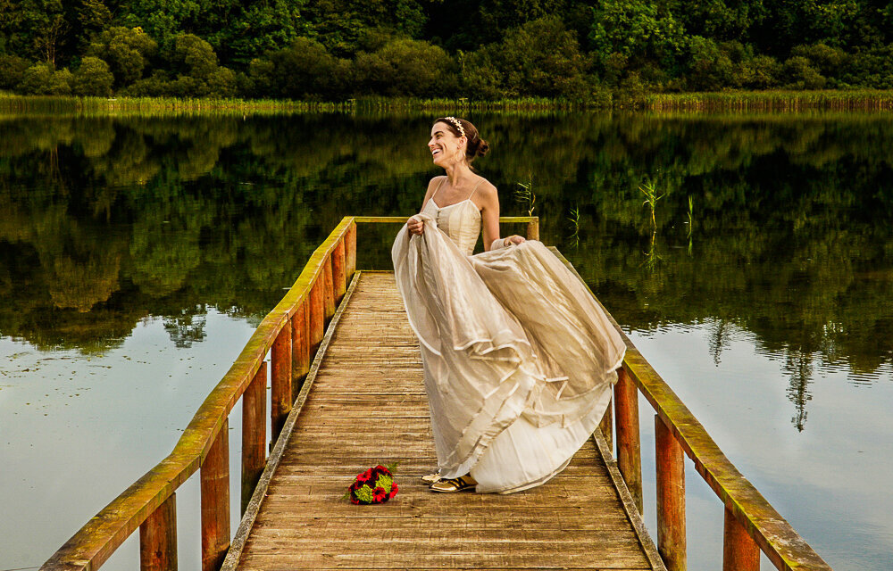 Bride with dark hair, wearing a leaf tiara and gold, boho style wedding dress and Gold Adidas trainers with colourful bouquet of flowers, while standing on a wooden jetty overlooking the lake at Castle Leslie Estate