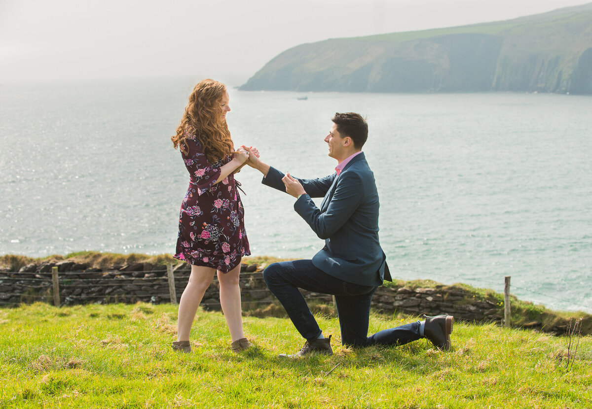 Young man going down on one knee to propose to his girlfriend in a field overlooking the sea