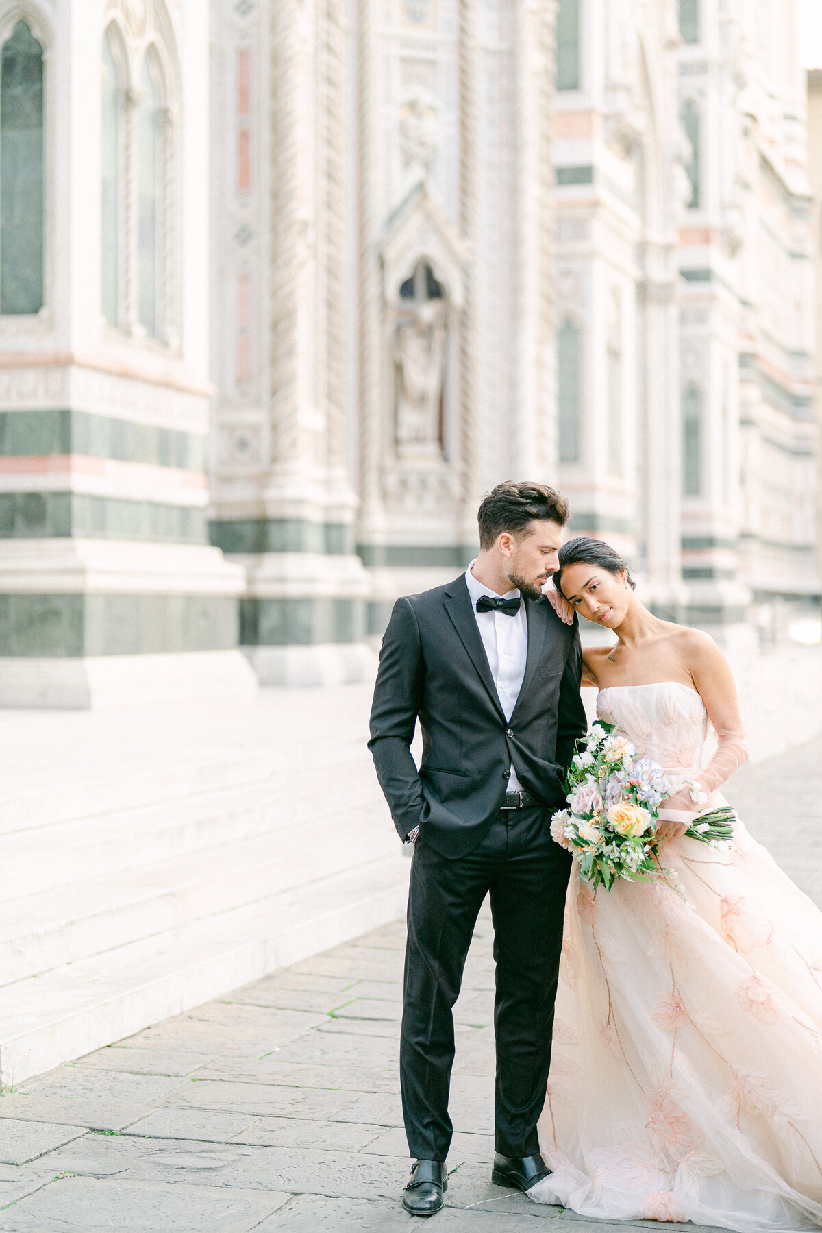 a bride resting her head on her grooms shoulder with a bouquet of flowers in her hands as they stand in front of the duomo in florence italy