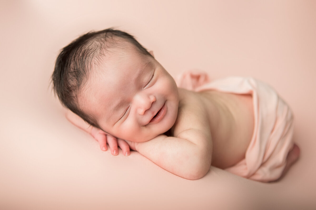 Smiling chin on hands newborn studio table pose on a pink backdrop