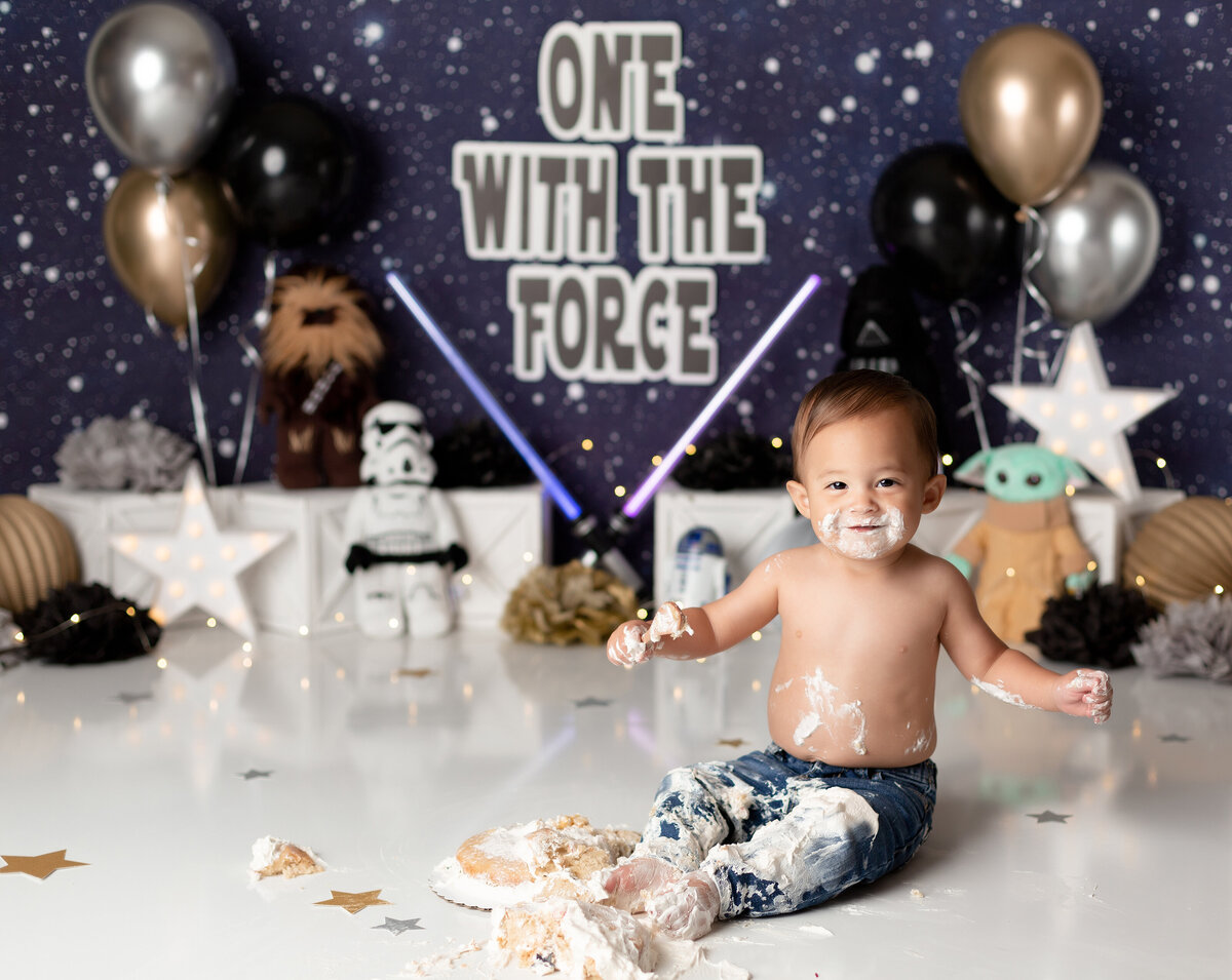 Star Wars themed cake smash in West Palm Beach and Boca Raton, FL photography studio. Baby is wearing jeans and has cake covering is pants and face. He is smiling at the camera. In the background there is a star backdrop, lightsabers, baby Yoda, Chewbacca, and a Stormtrooper.