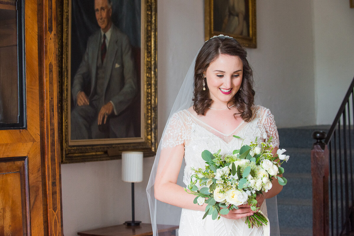 brunette bride wearing a vintage, empire style wedding dress looking at her white flower bouquet in the hallway of Westcove House, Kerry