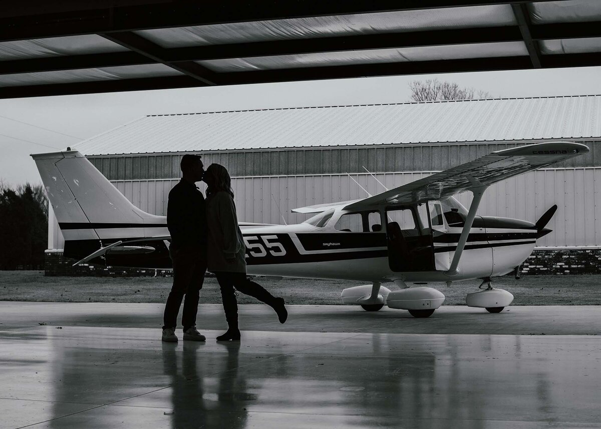 Maddie Rae Photography black and white silhouette of a couple kissing, she has her foot popped up behind her. they are standing in front of a small airplane