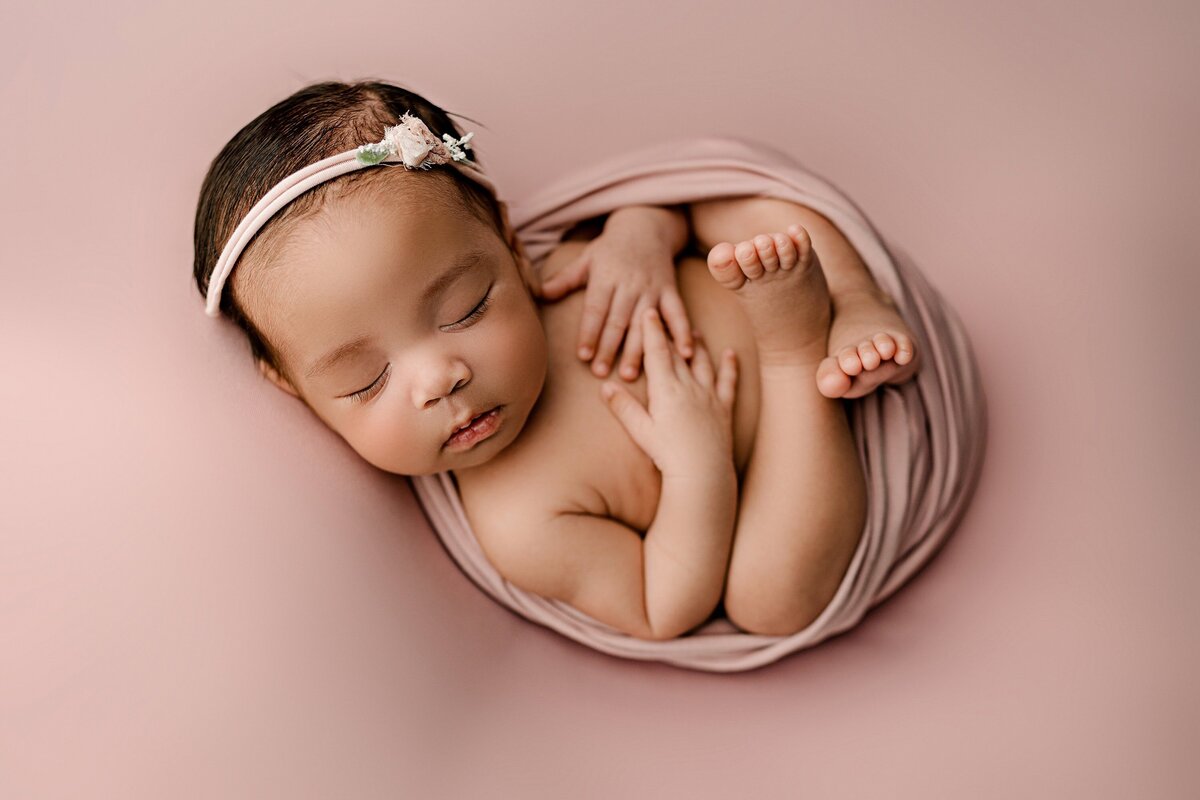 little newborn girl is curled up in a pink wrap wearing a matching bow in this  Charlotte studio