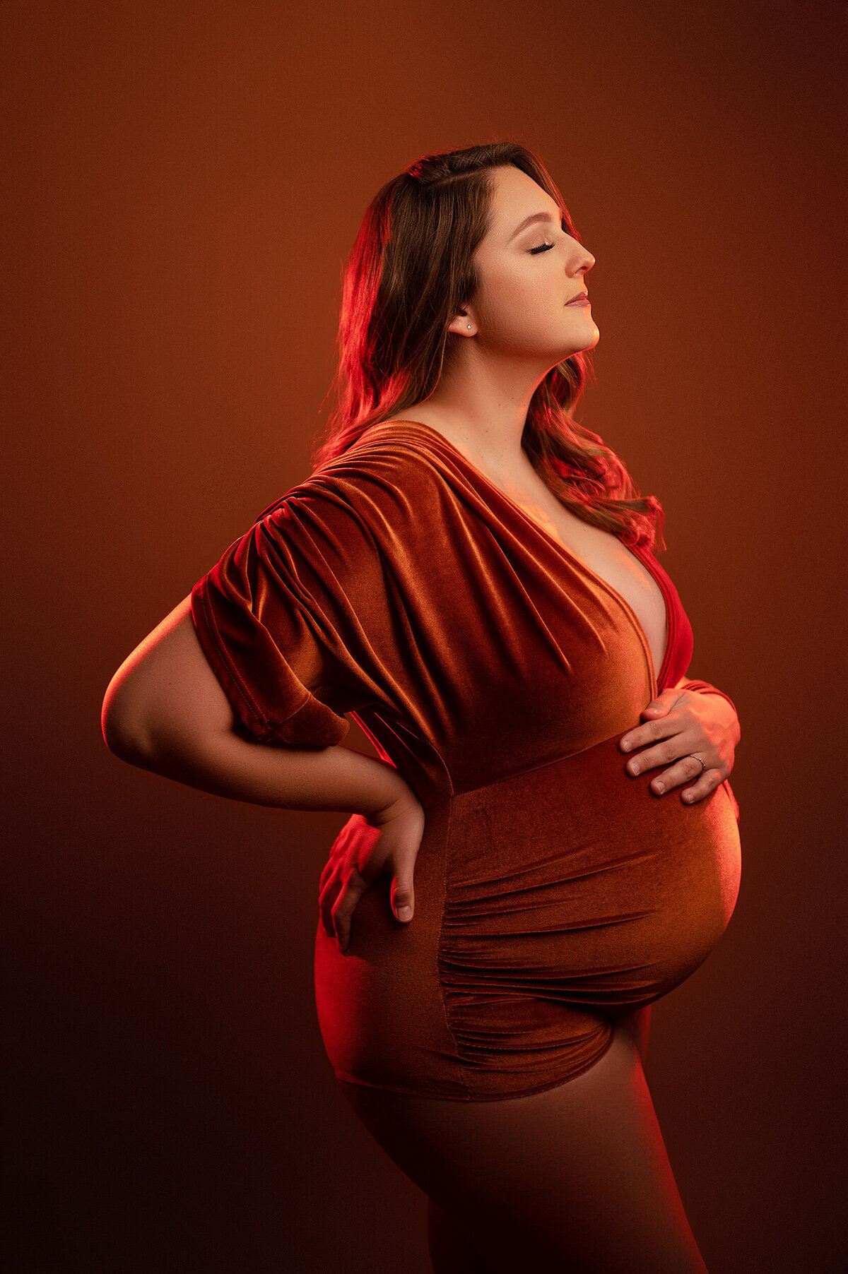 Profile shot of a pregnant woman wearing a plunging, red velvet romper framed in red studio light.