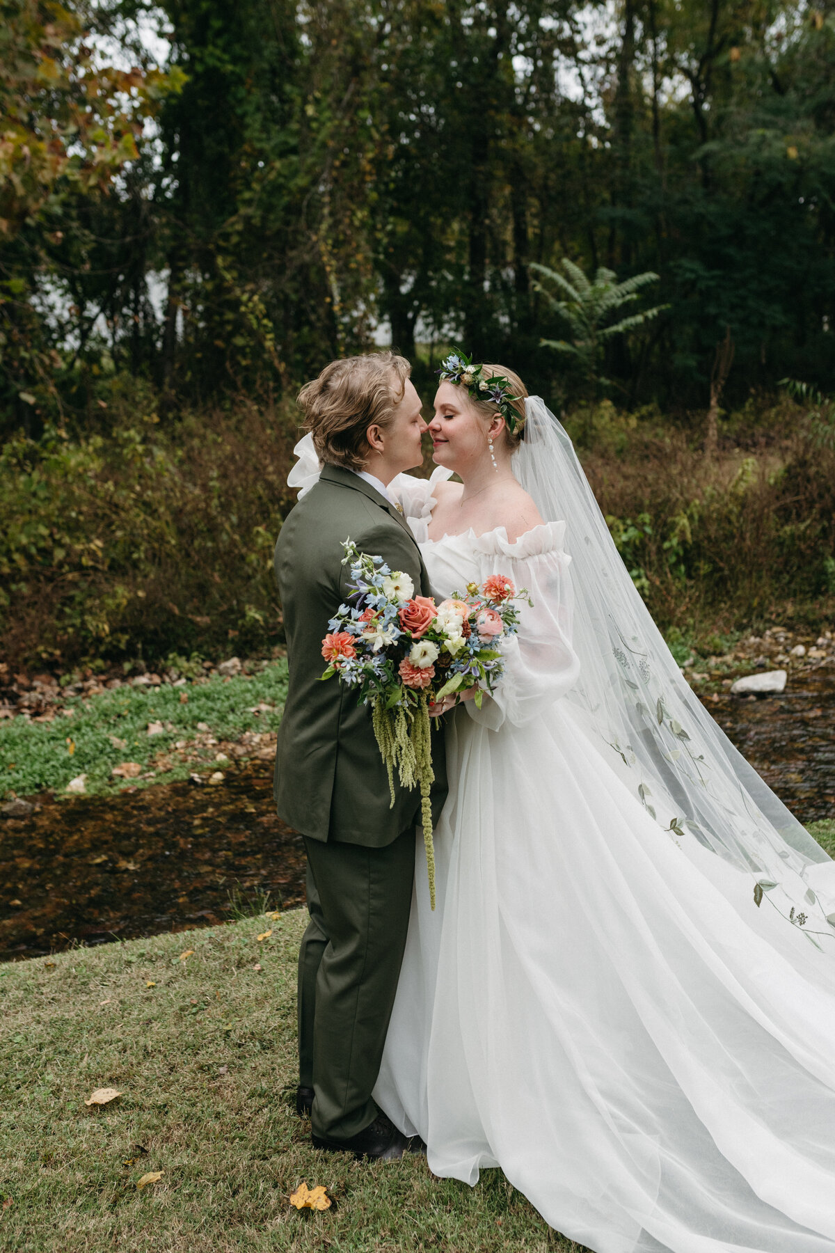 Bride and groom embracing beside a woodland creek, showcasing the bride’s flowing embroidered veil and her lush, colorful garden-style bouquet created by a fine art wedding florist in Northwest Arkansas.