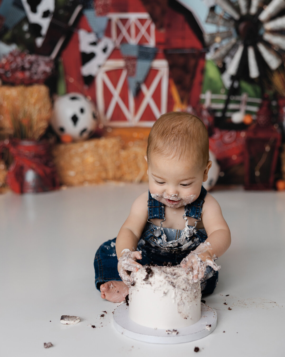 Baby celebrating his first birthday with a cake smash in front of a barnyard backdrop, smiling with frosting-covered hands. Photograph by Yucaipa cake smash photographer Kaitlyn Dawn Photography.
