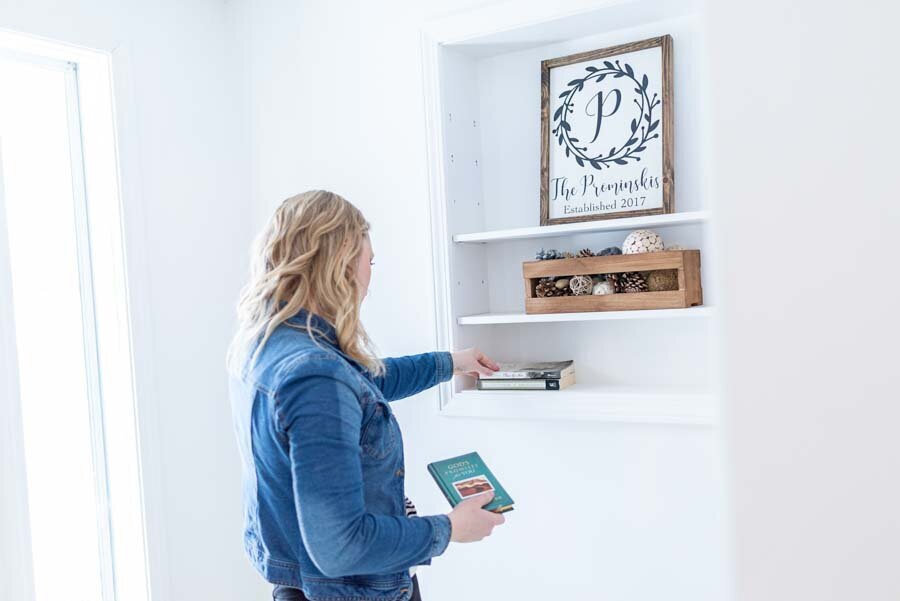 A woman in a denim jacket places a book on a white shelf, beside a framed picture and decorative items.