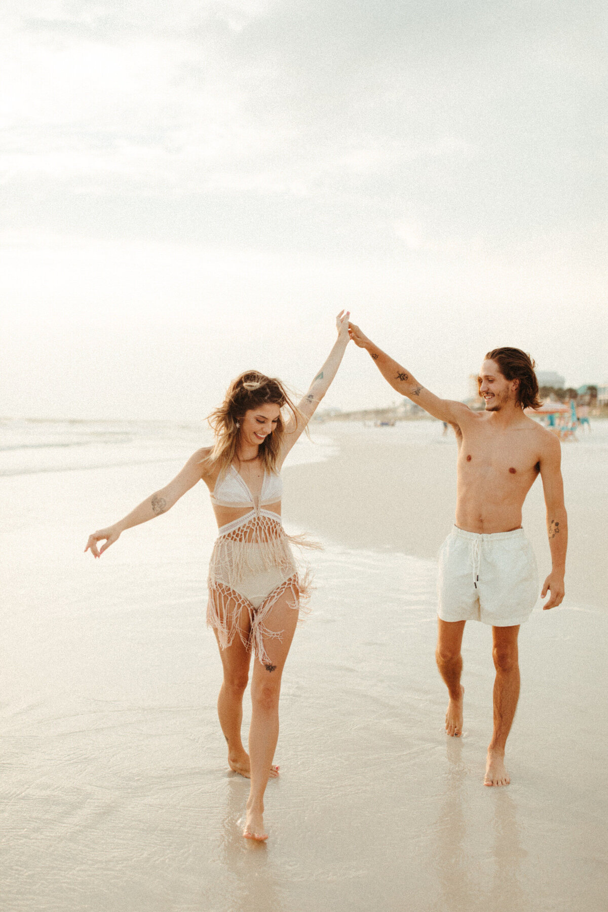 A guy in white swim trunks is walking along the beach and twirling a girl in a boho fringe swimsuit.