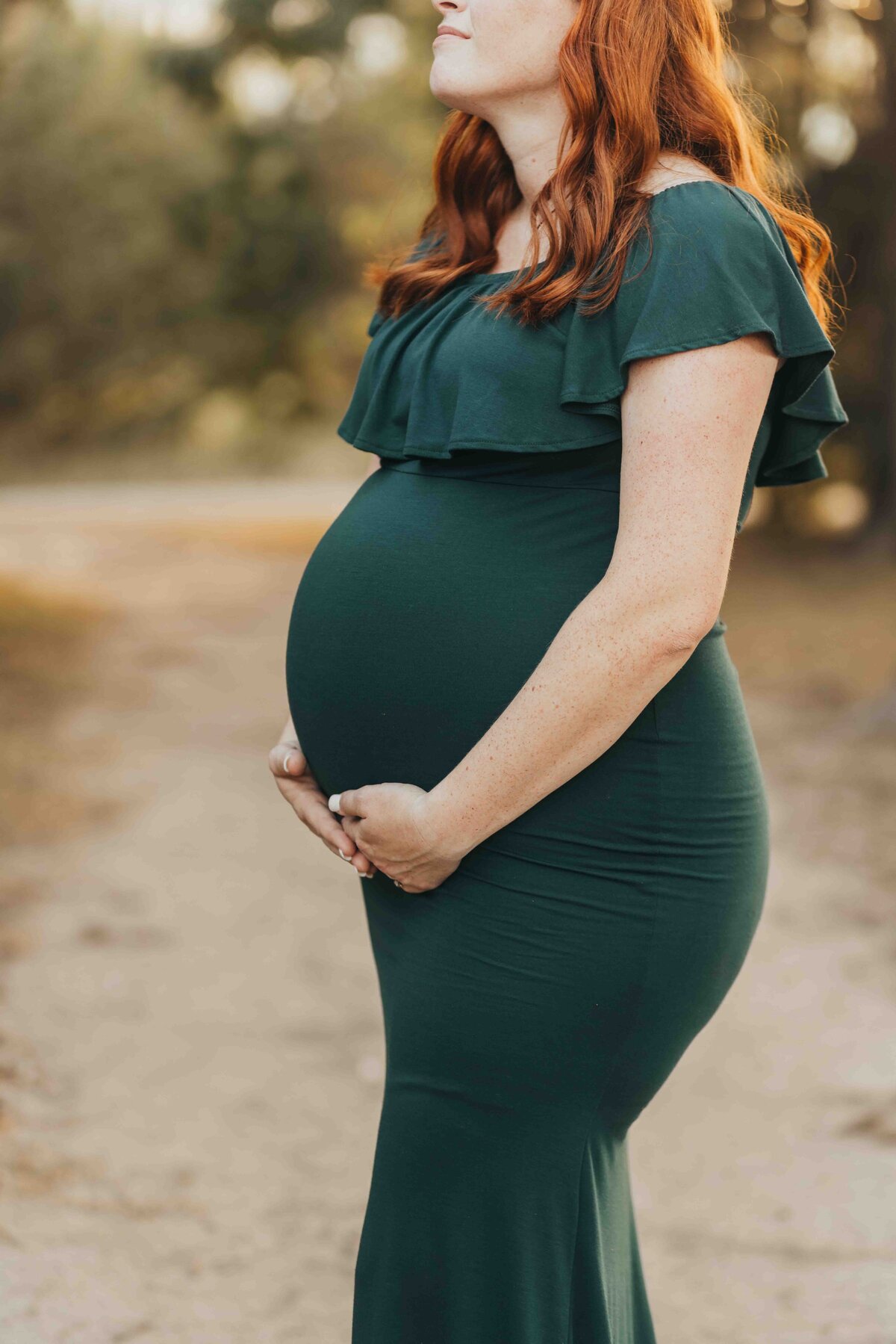 woman with red hair holds her belly, wearing a hunter green maternity gown.