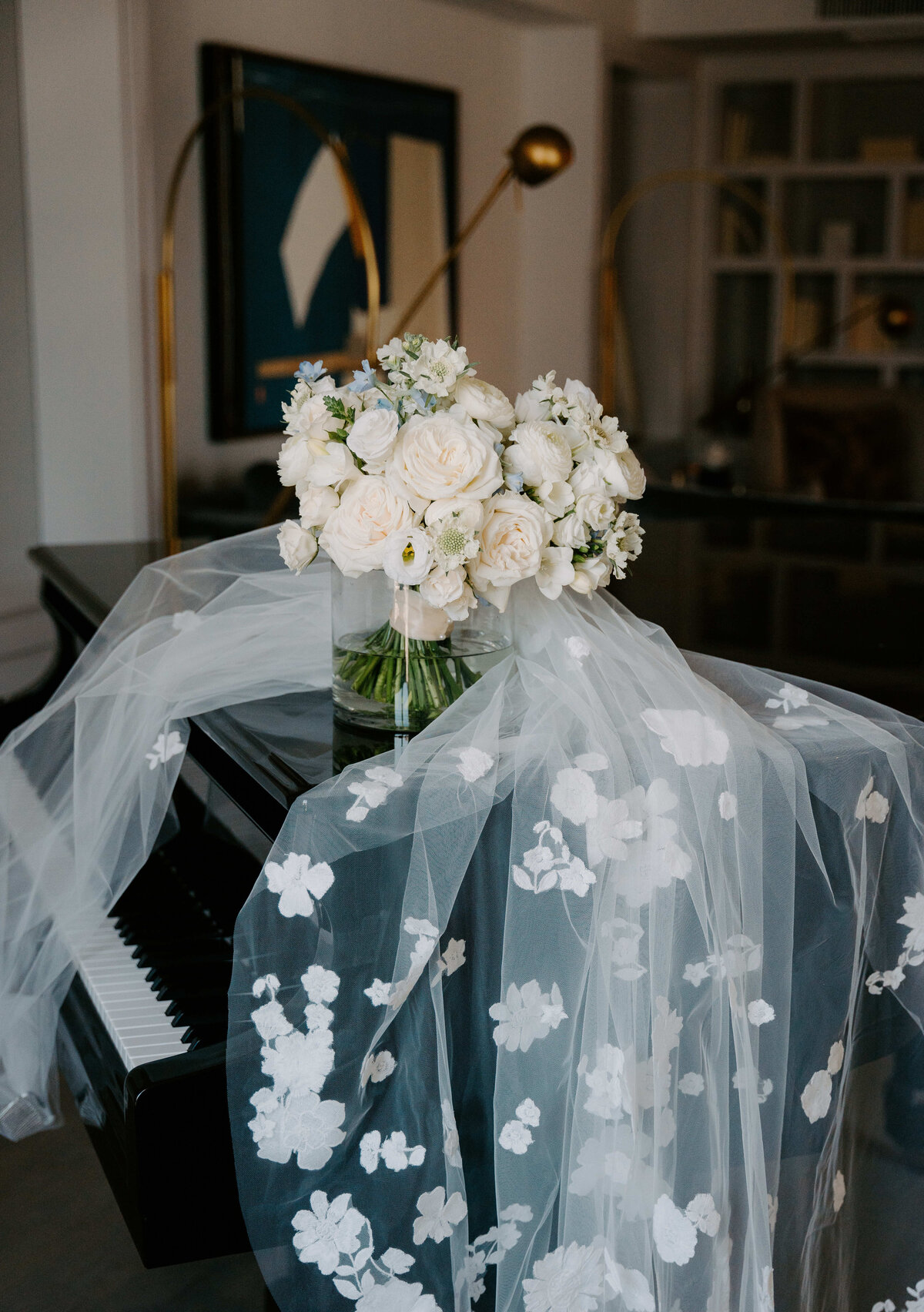A bouquet of white roses and flowers rests on a grand piano, draped with an embroidered floral veil. The setting is elegant and serene.