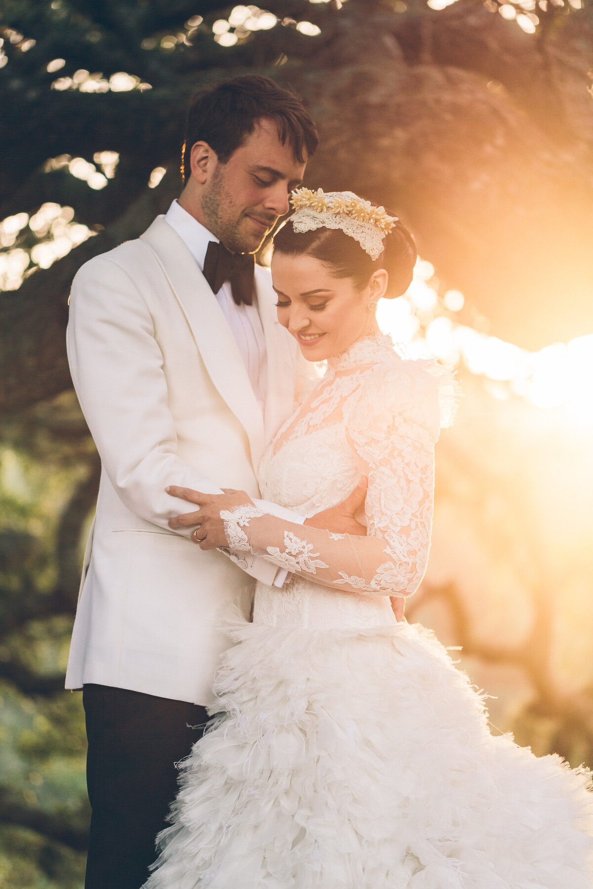 A photograph in color of Emma and John on their wedding day at Hearst Ranch in San Simeon, California. The bride and groom are in front of an old oak tree with the sun glowing from behind them. They are facing each other, the groom’s arms on her waist and the bride’s hand on his forearm. The groom is dressed in a white tuxedo jacket, black tie and black pants and he’s looking down at the bride. The bride is wearing an Alexander McQueen wedding dress with a bodice of lace with long sleeves and a long neckline along with a flowing skirt made with feathers. She wears a crown of lace and pearls. She is looking down and smiling. Wedding photography by Stacie McChesney/Vitae Weddings.