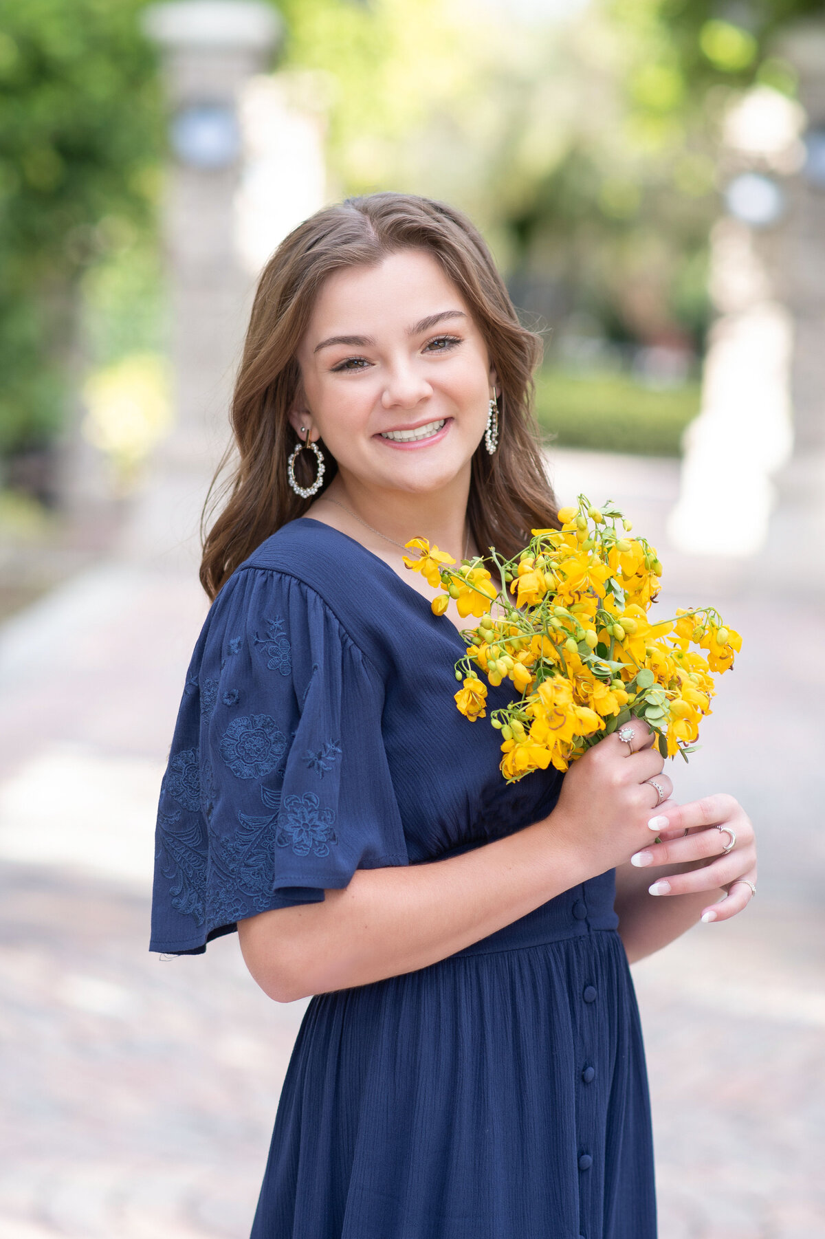 High school senior girl wearing a blue dress holing yellow flowers smiles at camera.