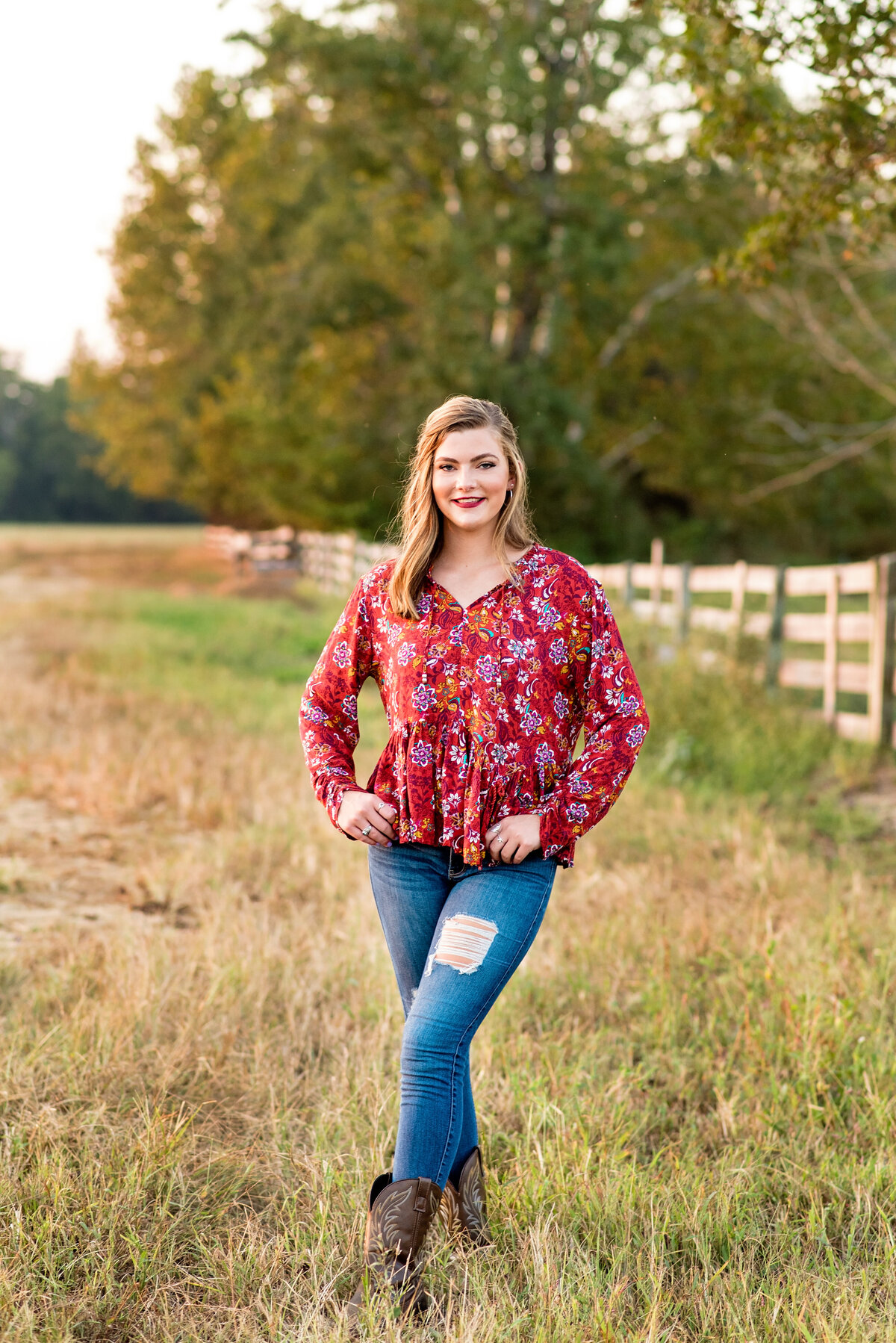 Senior girl poses in field in the country during sunset wearing jeans and a red blouse.
