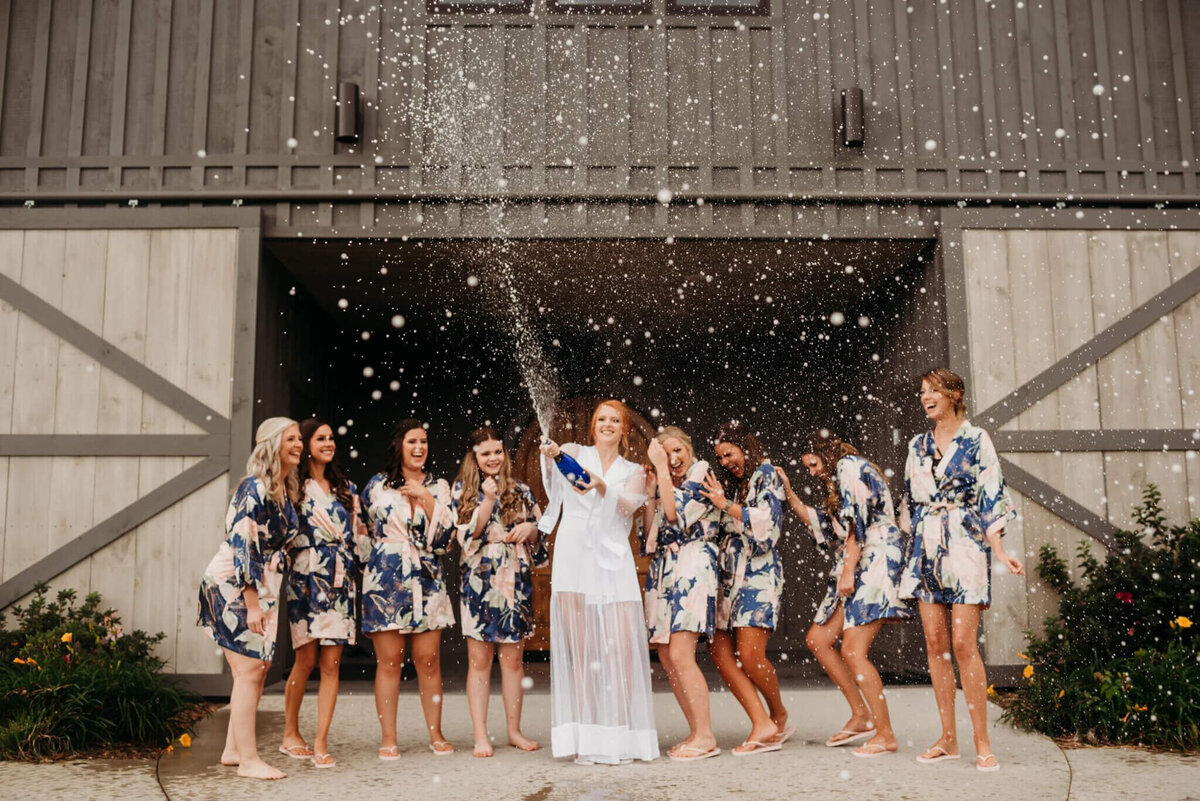 Photo of a bride in a white row popping champagne and smiling while her bridesmaids are cheering in navy robes and a gray barn in the background