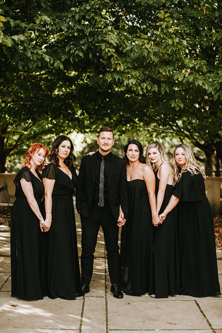 The wedding party poses outside wearing black gowns and a black suit, with large trees in the background.