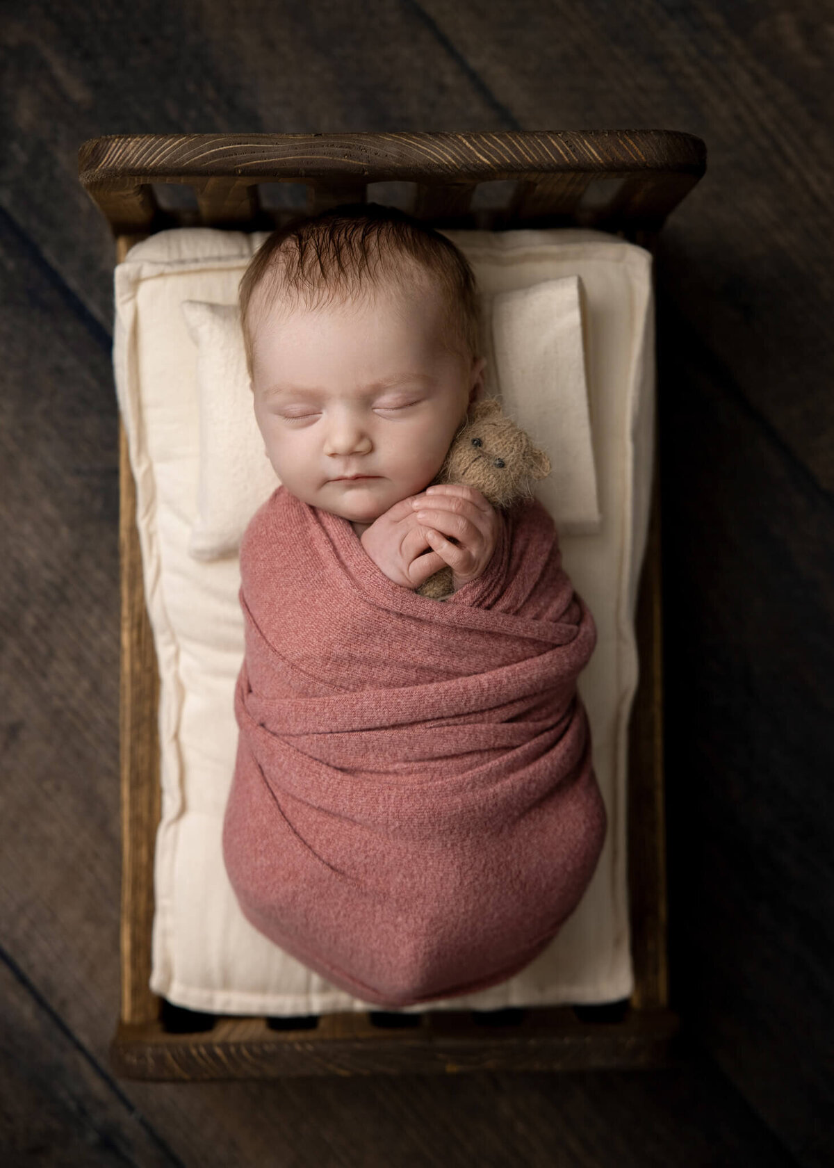 newborn baby wrapped in a pink fabric asleep in a tiny bed holding a tiny teddy bear