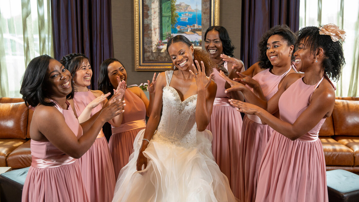 A fun getting ready photo where  a bride is showing her engagement ring to her bridesmaids wearing pink dresses.