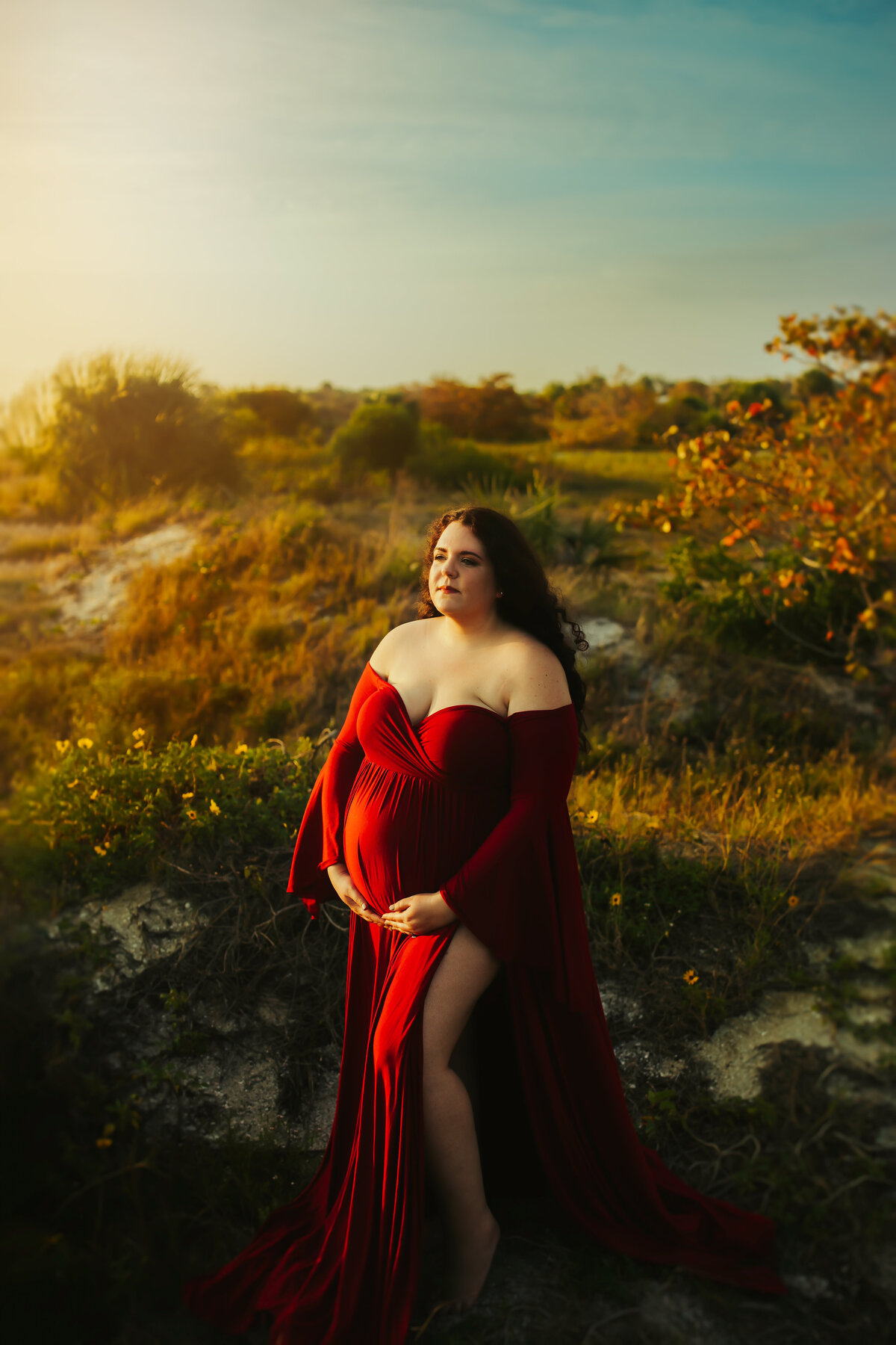 colorful portraits of a pregnant women in a red off shoulder gown facing the sun at the beach