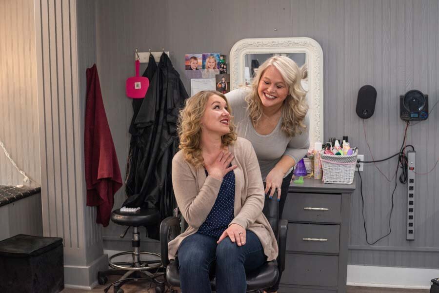 Two women smiling in a salon, one sitting in a stylist's chair, with hair accessories and a mirror in the background.